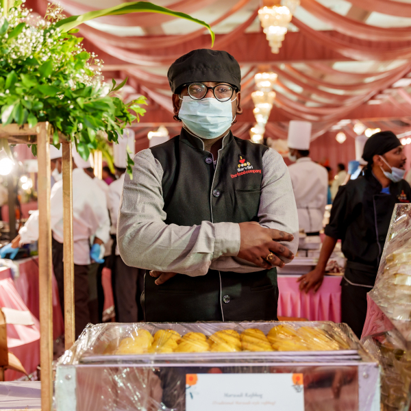 A catering staff member in a black uniform and face mask stands behind a dessert display at an elegant event, with other staff and a decorated venue in the background.